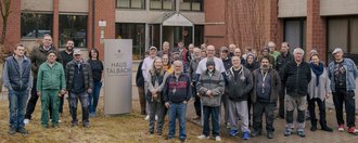 Haus Talbach Vor dem Haupteingang stehen Bewohnende und Mitarbeitende von Haus Talbach und lächeln in die Kamera.
