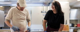 Haus Talbach Zwei lachende Menschen arbeiten gemeinsam in der Küche.