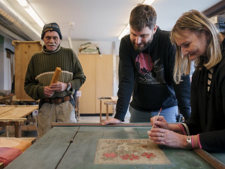 Eine Person bemalt eine Holztür in einer Schreinerwerkstatt, während zwei andere Personen interessiert dabei zusehen.